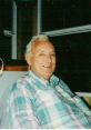 Smiling elderly man wearing a plaid shirt, seated indoors, exuding warmth and friendliness in a cozy setting.