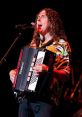 Weird Al" Yankovic passionately performs with an accordion, wearing a colorful shirt against a vibrant stage backdrop.