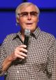 Adam West speaking at an event, wearing sunglasses and a plaid shirt, holding a microphone, showcasing his iconic charm.