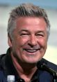 Alec Baldwin smiles brightly at a speaking event, showcasing his charismatic personality and signature hairstyle.