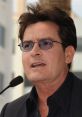 Charlie Sheen speaking at a public event, wearing glasses and a black suit, showcasing his unique style and persona.