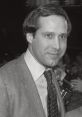 Chevy Chase smiling while wearing a suit and tie, surrounded by fans and media in a black-and-white scene.