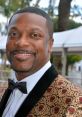 Chris Tucker smiles at an event, sporting a stylish black and gold patterned suit, showcasing his charismatic personality.