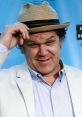 John C. Reilly tipping his hat, dressed in a light suit, smiling against a blue background at a film event.