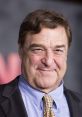 John Goodman smiling confidently at a press event, showcasing his signature charm and style in a suit and tie.