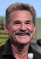 Kurt Russell smiling at a panel discussion, showcasing his iconic style and charm in a casual black shirt.