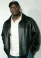 Michael Clarke Duncan smiles confidently in a black leather jacket and cap, showcasing his iconic style and charm.