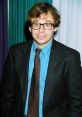 Rick Moranis smiling in a stylish outfit, featuring a blazer, blue shirt, and patterned tie, highlights his iconic 90s look.