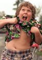 Chunk from The Goonies, laughing joyfully, wears a colorful floral shirt and plaid shorts in an energetic moment.