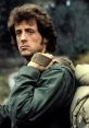 John J. Rambo with an intense expression, wearing military gear and carrying a heavy backpack in a rugged outdoor setting.