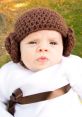 Cute baby dressed as Princess Leia in a cozy white outfit with a brown belt and iconic brown earmuff hat.