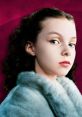 Veruca Salt poses confidently with striking blue eyes and a fur coat against a vibrant pink background.