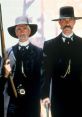 Two men in dark attire and hats pose with weapons, embodying iconic characters from *Tombstone (1993)*.