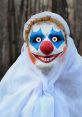 Eerie clown with bright face paint and red nose, shrouded in white, surrounded by dark trees in a spooky setting.