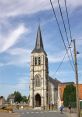 Historic church with a tall spire in a quaint French village, surrounded by greenery and a peaceful street scene.