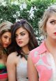 Three young women pose with a background of white flowers, showcasing vibrant fashion and a sense of intrigue.