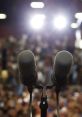 Microphones in focus with bright lights, capturing the anticipation in a crowded audience before an important speech.