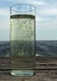 Tall glass of water with green particles, set against a serene ocean backdrop on a wooden surface.