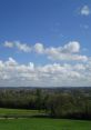 Vibrant green fields under a clear blue sky with fluffy clouds, showcasing a peaceful countryside scene and pleasant weather.