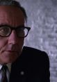 Warden from Shawshank Redemption, wearing glasses, with a serious expression in a stark, gray setting.