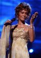 Whitney Houston performing passionately in a glamorous gold gown, showcasing her iconic vocal talent on stage.