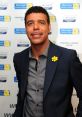 Chris Kamara smiling at a charity event, wearing a dark blazer and a yellow flower pin, promoting cancer awareness.