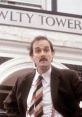 Basil Fawlty stands in front of Fawlty Towers, showcasing his iconic comedic expression and classic striped tie.