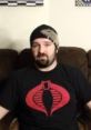 Darksydephil wearing a Cobra logo shirt, seated on a couch with artwork in the background, exudes a casual gaming vibe.