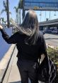 Long-haired individual wearing black, walking down a sunny street with palm trees and a bag, embodying a relaxed vibe.