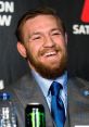 Conor McGregor smiles confidently during a press conference, showcasing his stylish suit and iconic beard.