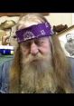 Man with long beard and purple bandana, seated in a cluttered room, reflecting a unique and unconventional style.