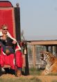 Joe Exotic sits on a lavish throne, adorned in a crown and robe, beside a roaring tiger in a wildlife sanctuary setting.