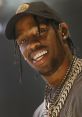 Travis Scott grinning on stage, showcasing his signature style with chains, a cap, and sparkling grills in a vibrant performance.