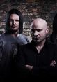 Disturbed band members pose against a textured brick wall, showcasing their iconic rock aesthetic and intense energy.