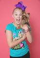 Joyful girl with a large purple bow, wearing a Jojo-themed shirt, holds a small dog against a bright pink background.