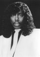 Rick James in a stylish white suit with a black tie, showcasing his iconic curly hairstyle and captivating expression.