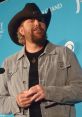 Toby Keith, country music star, speaks at a press event wearing a cowboy hat and denim jacket. Engaging personality shines through.