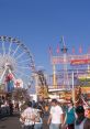 Visitors enjoy lively fairgrounds with a Ferris wheel, rides, and vibrant attractions under a clear blue sky.