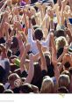 Crowd of enthusiastic fans cheering outdoors with hands raised, celebrating an exciting live event under the sun.