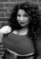 Confident woman with curly hair poses against a brick wall, showcasing bold style and self-expression in black and white.