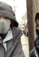 Person wearing a gray mask and coat sits beside a dog on a city street, with cars parked in the background.