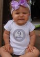 Nine-month-old baby girl in a white onesie with a "9 Months" badge, wearing a purple bow, smiling happily.