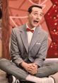 Pee Wee Herman in a playful pose, wearing a gray suit and red bow tie, surrounded by colorful props and a whimsical backdrop.