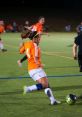 Female soccer player in an orange jersey dribbles the ball on a green field during a night match, showcasing football skills.