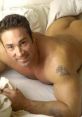 Smiling man with a muscular build lying on a bed, showcasing a tattoo and exuding confidence, embodying Gachi Pack charm.