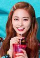 Brightly smiling individual holding a pink drink, showcasing a vibrant, colorful outfit against a turquoise backdrop.