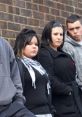 Group of young individuals with serious expressions, standing against a brick wall, embodying a gang-like aesthetic.