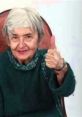 Elderly woman in a green sweater giving a thumbs-up, radiating positivity and warmth, representing Stilwell Grandma 2 spirit.