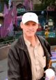 Chris The Hacker smiling at a vibrant amusement park, wearing a cap and holding a drink, surrounded by colorful attractions.