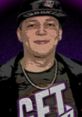 MontanaBlack sporting a cap and chain, featuring a bold graphic shirt in vibrant colors, set against a purple background.